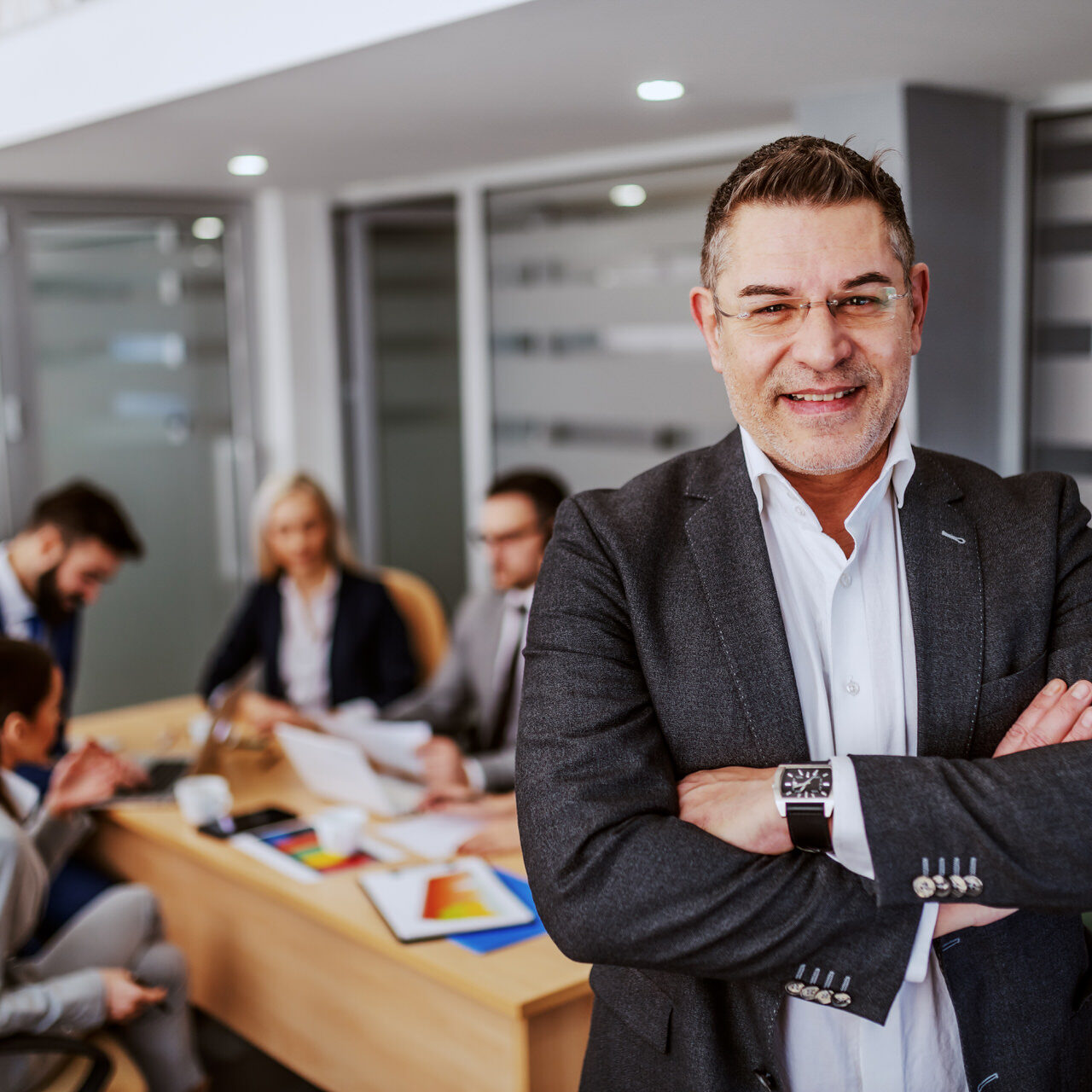 Middle aged successful caucasian CEO standing in boardroom with hands crossed and looking at camera. In background are his employees working on project for important client. Middle aged successful caucasian CEO standing in boardroom with hands crossed and looking at camera. In background are his employees working on project for important client.